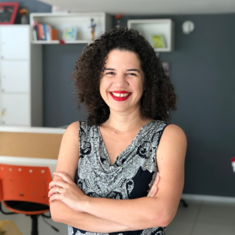 Foto de Eugenia olhando para a câmera de braços cruzados na frente no corpo, cabelo solto e sorriso no rosto