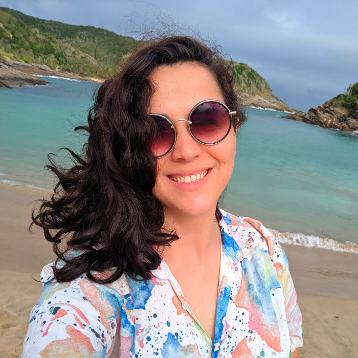 Foto de mulher branca com cabelos soltos para o lado, sorrindo, usando óculos de sol. Ela está na praia.